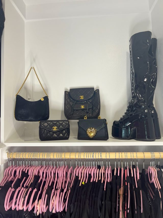 A shelf of black designer handbags and patent boots above a row of hanging pink clothing