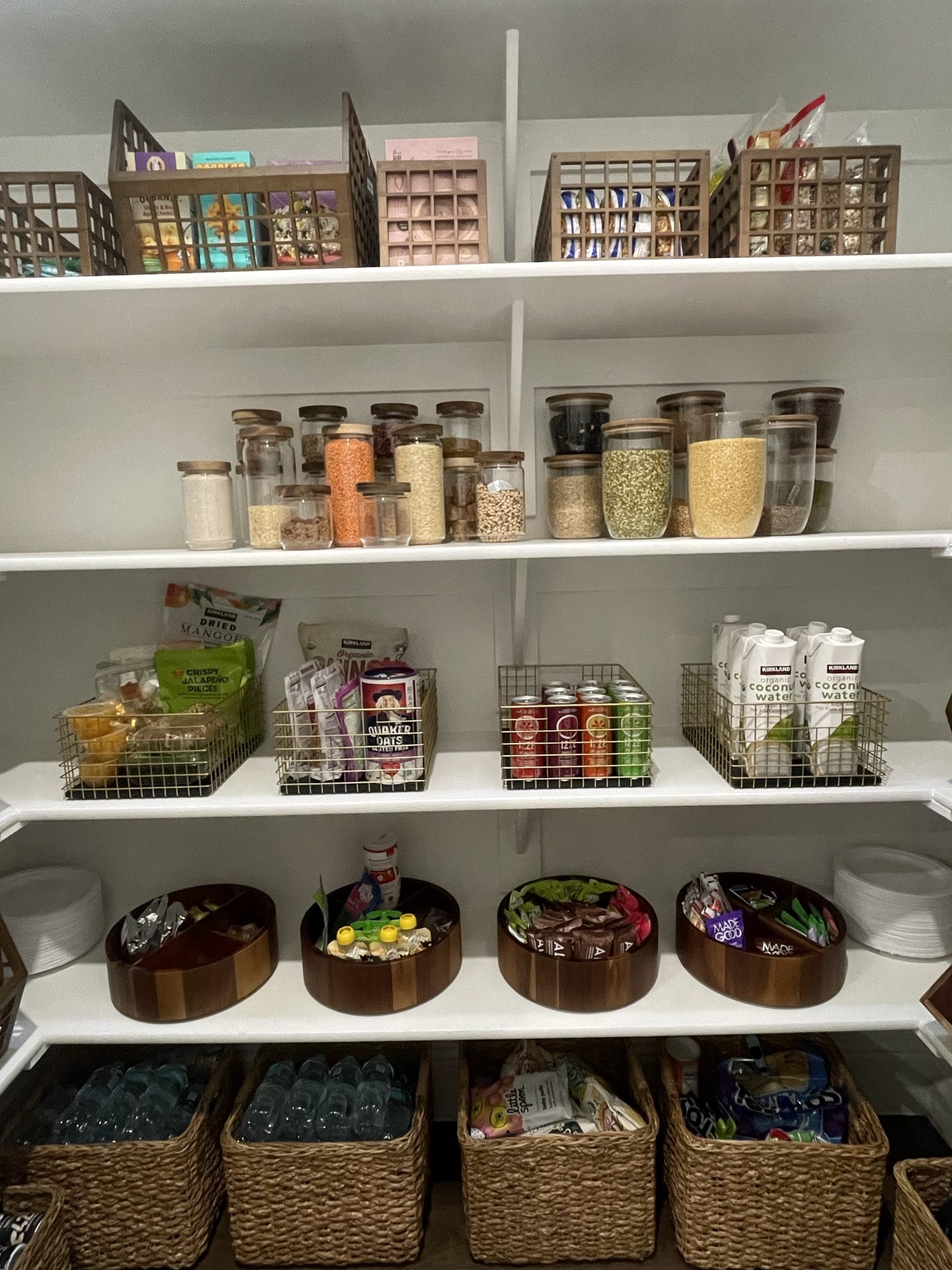 A pantry with dry goods decanted into glass jars, wire baskets of drinks and snacks, and woven baskets on the lower shelf