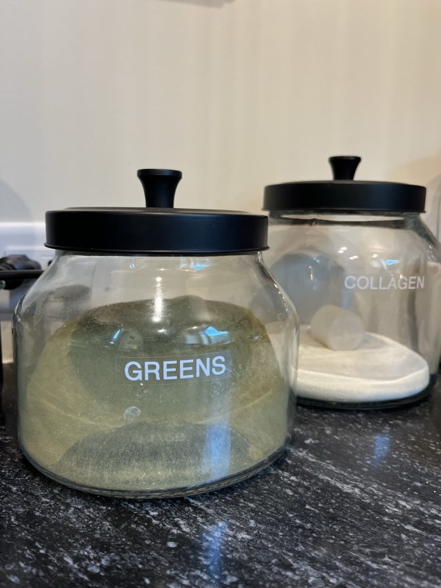 Labeled glass canisters of greens powder and collagen on a dark counter